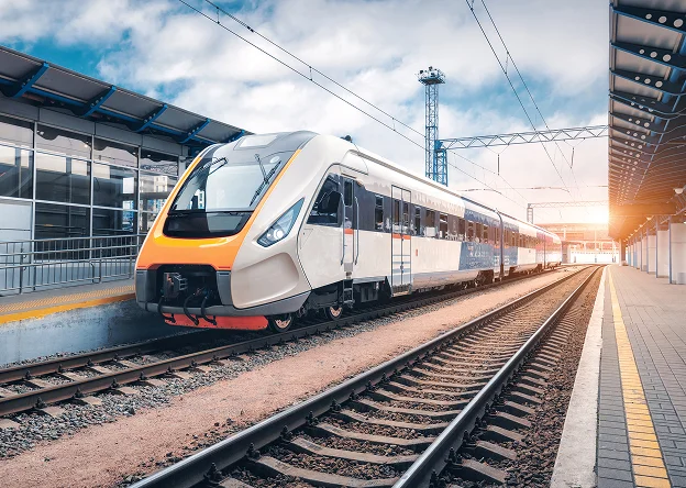 Un train moderne et orange-blanc arrive à une gare ensoleillée sous un ciel nuageux.