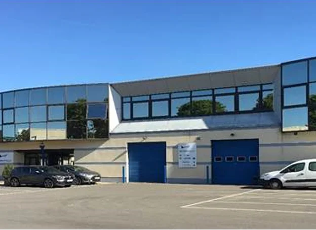 Bâtiment moderne avec de grandes baies vitrées bleues, deux portes de garage et des voitures garées.